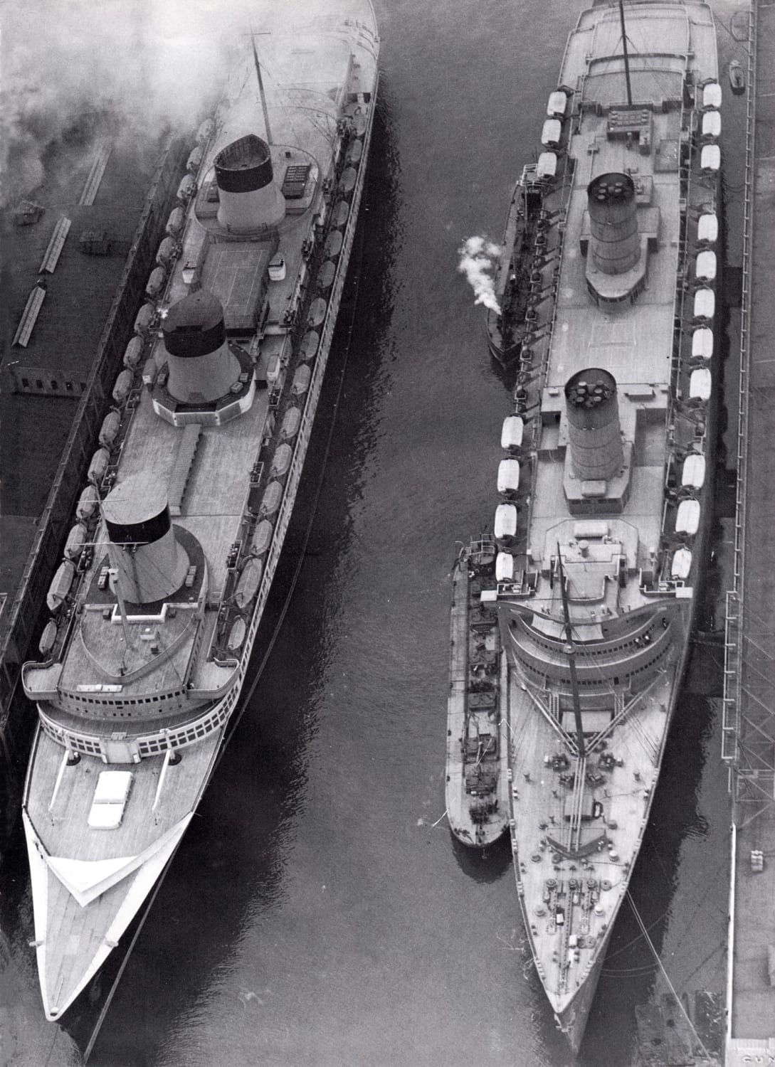 British Ocean Liner RMS Queen Elizabeth in her wartime grey colors on right and French Ocean Liner SS Normandie on left in New York City, circa 1941. QE would survive the War, while Normandie wouldn't leave that dock. A fire would claim her.