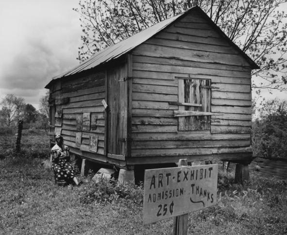 A Louisiana woman sitting outside a wooden shack, on the back of which some paintings are hung. A sign says "ART EXHIBIT ADMISSION 25¢ THANKS". 1952.