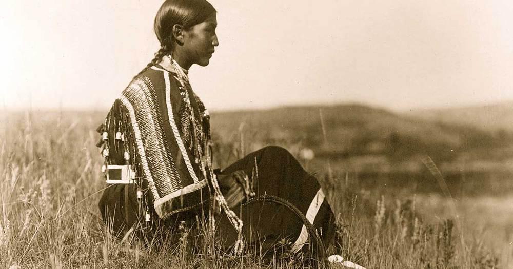A young Piegan woman sits on a hilltop wearing a traditional beaded dress. The photo dates to around 1910.