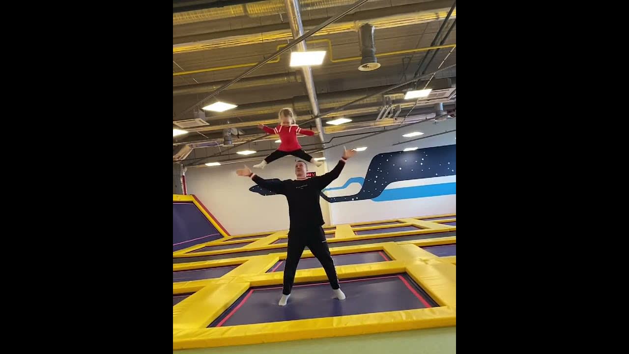 Dad Holds Toddler's Feet in Hands While Jumping on Trampoline in Jumping Park - 1200087