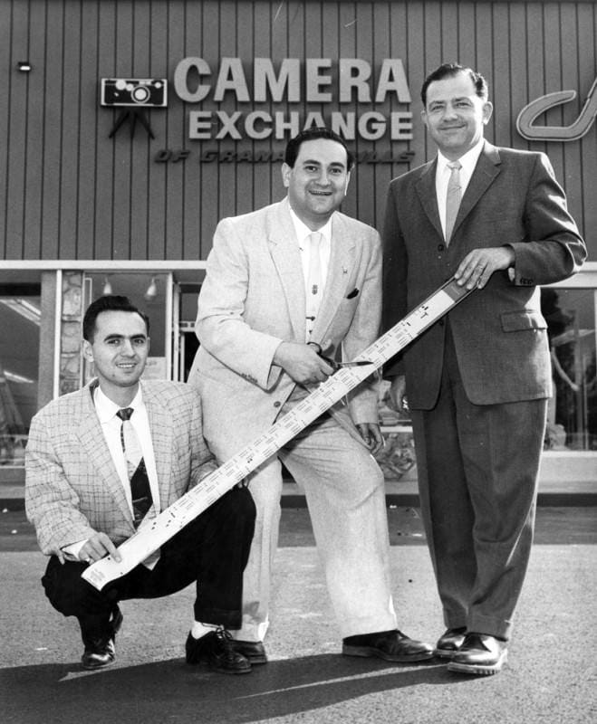 Photograph caption dated March 5, 1959 reads, "Owners of Camera Exchange of Granada Hills (CA) use strip of film instead of conventional ribbon as they prepare to 'cut the ribbon' to open their fourth Valley store. "