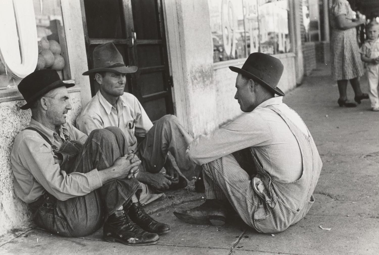 1939. Farmers sitting against wall and squatting on sidewalk, Spur, Texas. Photographer Lee Russell