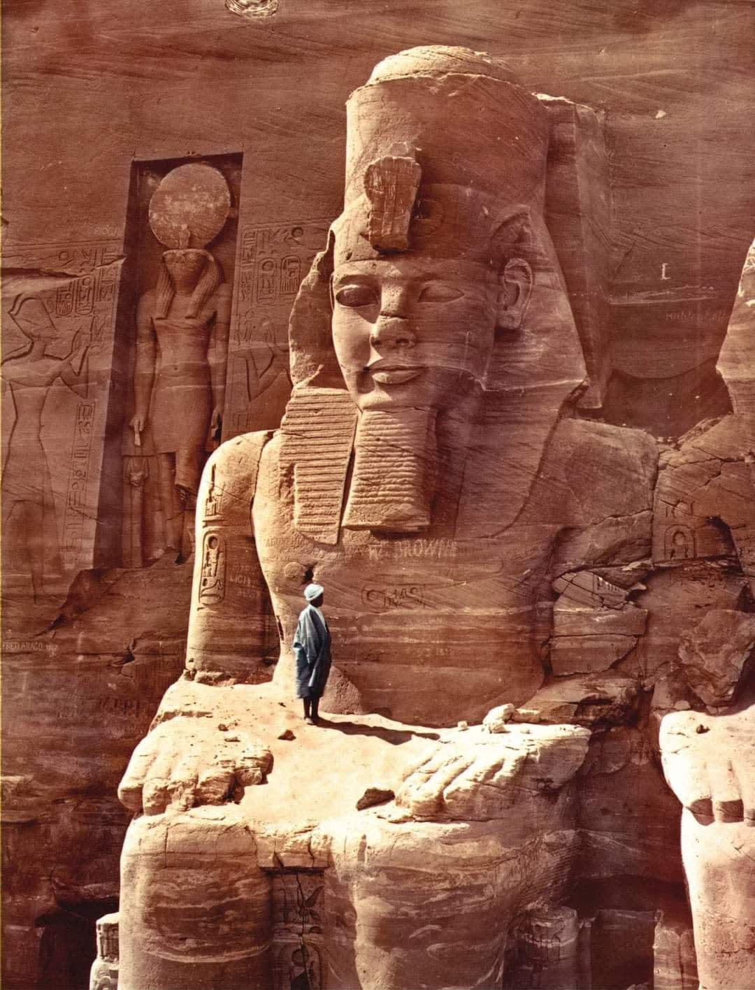 Colourised photo of a man standing on a colossal figure of Ramesses II at the temple of Abu Simbel, Egypt, 1865
