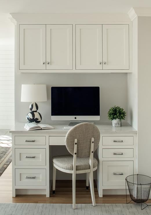 A gray round back chair sits at a white built-in kitchen desk boasting white drawers donned with polis… | Kitchen office nook, Home office space, Home office design