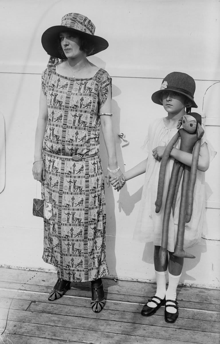 Socialite Mrs. Katherine Williamson and her nine-year-old daughter, Margaret, aboard the SS Paris as it arrives in New York from France. Katherine is wearing a fashionable new ‘Cleopatra’ gown, the current haute-couture. Margaret is carrying one of the new “daddy dolls.”