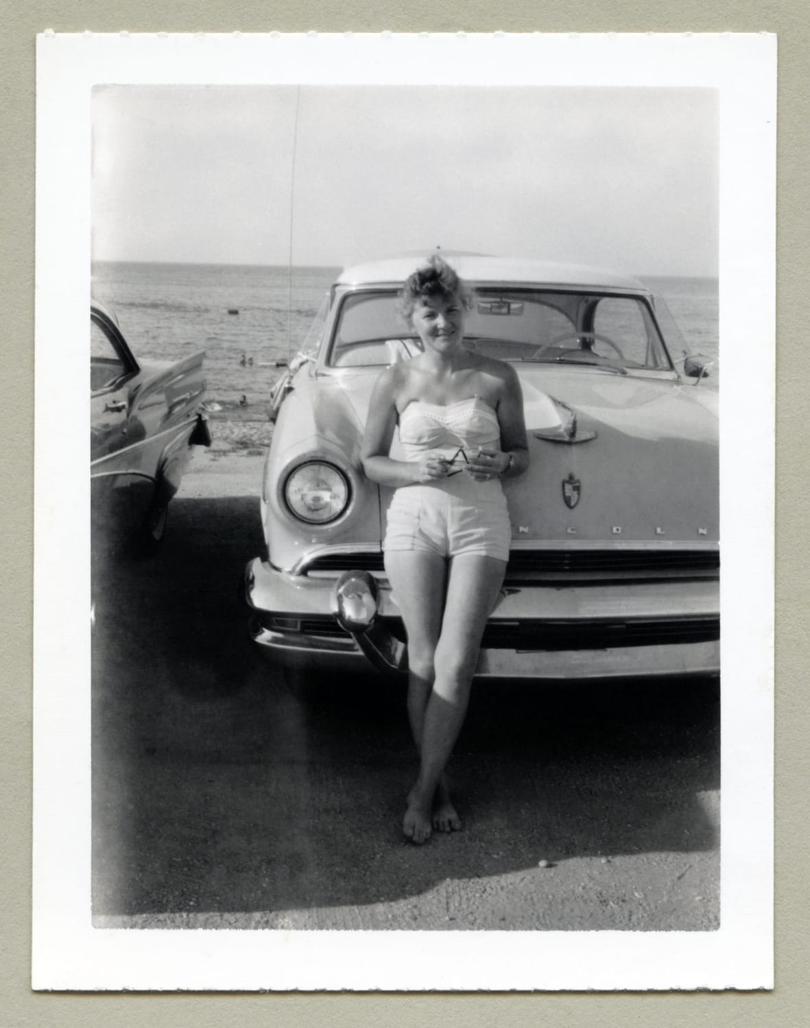 A cheerful lady posing with a 1955 Lincoln Capri on the beach. She's dressed in a strapless swimsuit in the fashion of the 1950s