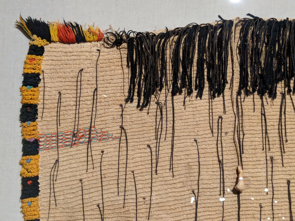 onview in Gallery 31: This Māori cloak in the 'korowai' style is decrorated with multicolored woolen elements and twisted black-dyed flax tags called 'hukahuka.'