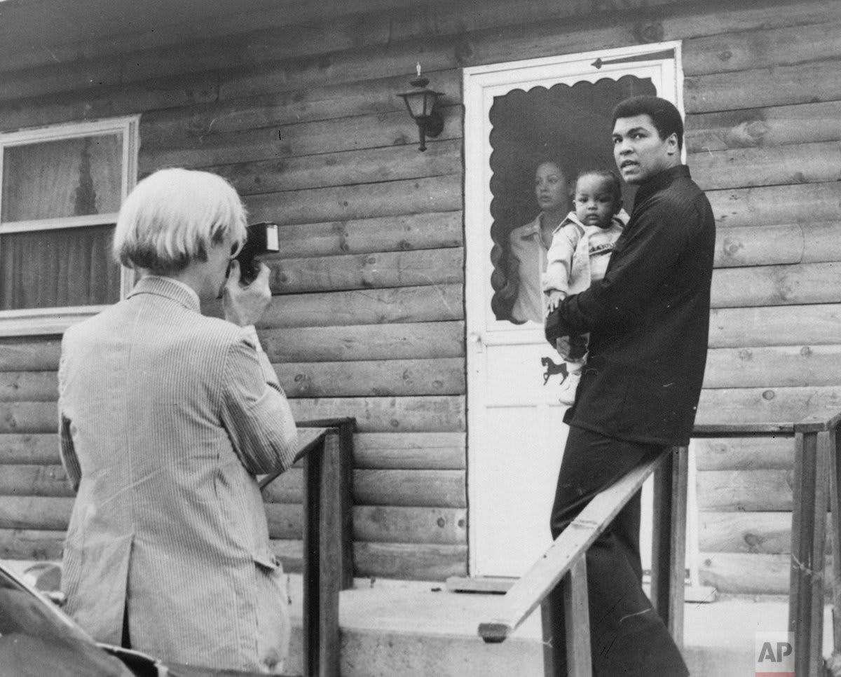 Pop artist Andy Warhol, left, is shown photographing Muhammad Ali, his infant daughter, Hanna, and wife, Veronica, at Ali's training camp in Deer Lake, Pa., OTD in 1977.