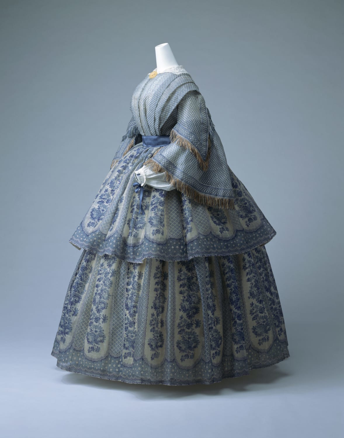 This 1855 day dress proves to be very fashionable for the time with various decorative elements such a fringe and bold patterns, reflecting the rise of voluminous garments and heavy embellishments that became so popular. Read more!