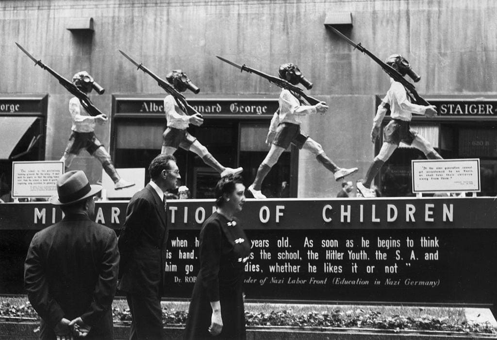 People view an anti-Nazi display with statues of young boys wearing uniforms, armbands, gas masks and carrying rifles. Rockefeller Center, New York City, 1945.