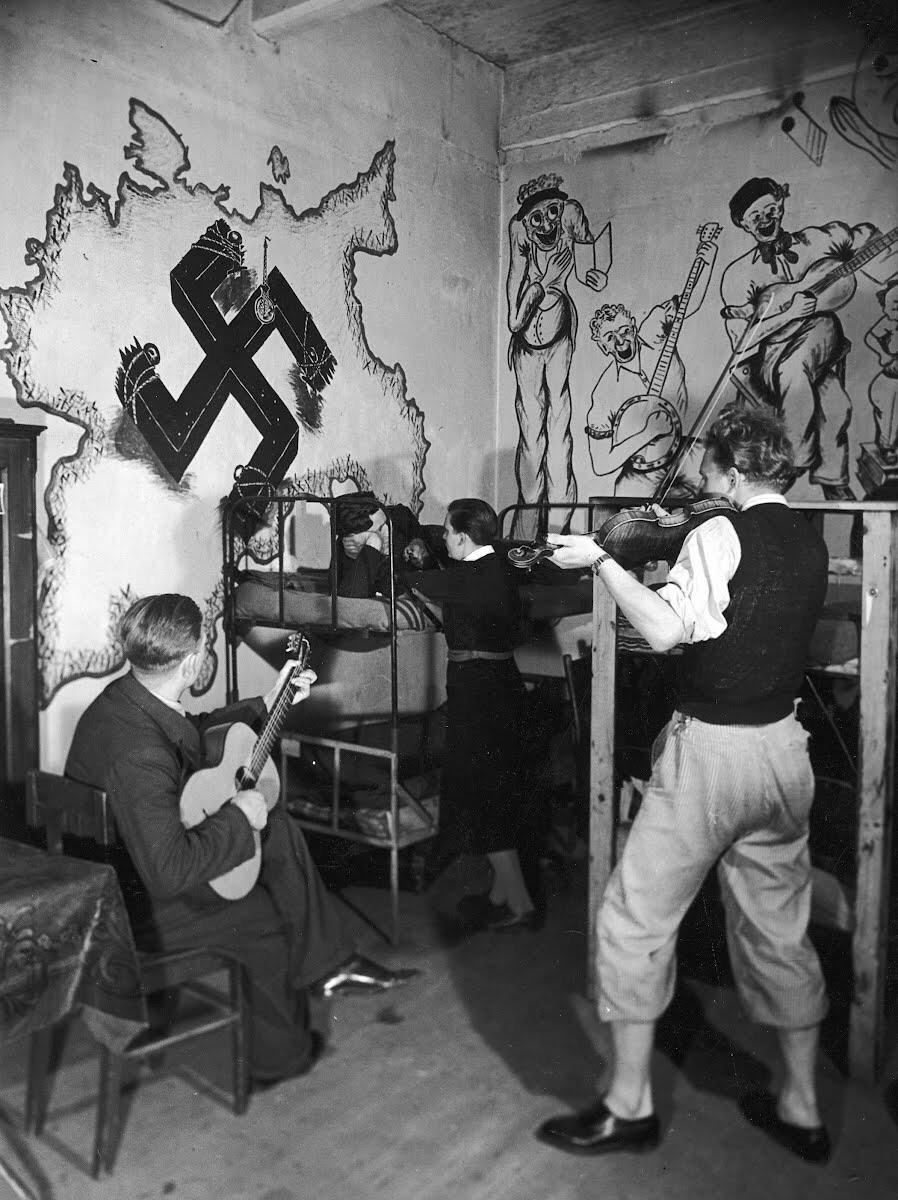 Jewish refugees from Nazi Germany playing violin and guitar as others chat in their bunks next to a German wall map emblazoned with a swastika in the cellar of an old abandoned factory they live in on the outskirts of Prague, Czechoslovakia, 1938.