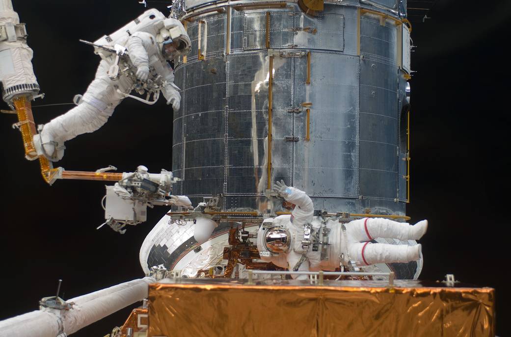 STS-125 Mission Specialist Andrew Feustel navigates near the Hubble Space Telescope on the end of the remote manipulator system arm, controlled from inside Atlantis' crew cabin. Mission Specialist John Grunsfeld signals to his crewmate from just a few feet away.