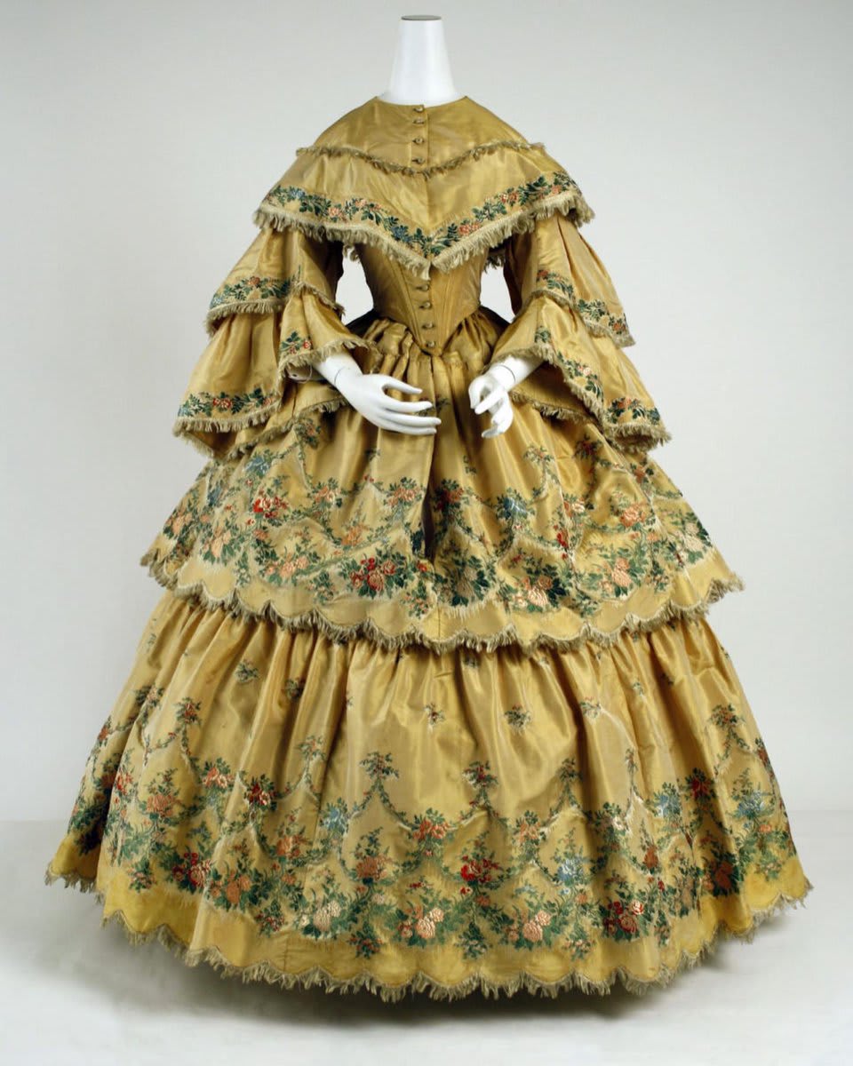 This 1859 ensemble has a gold, two-tiered skirt and pagoda sleeves, both of which are edged in a red, green, and peach floral brocade pattern. It has a round-necked bodice with a frayed v-shaped seam above the bust and frayed edges. Read more!